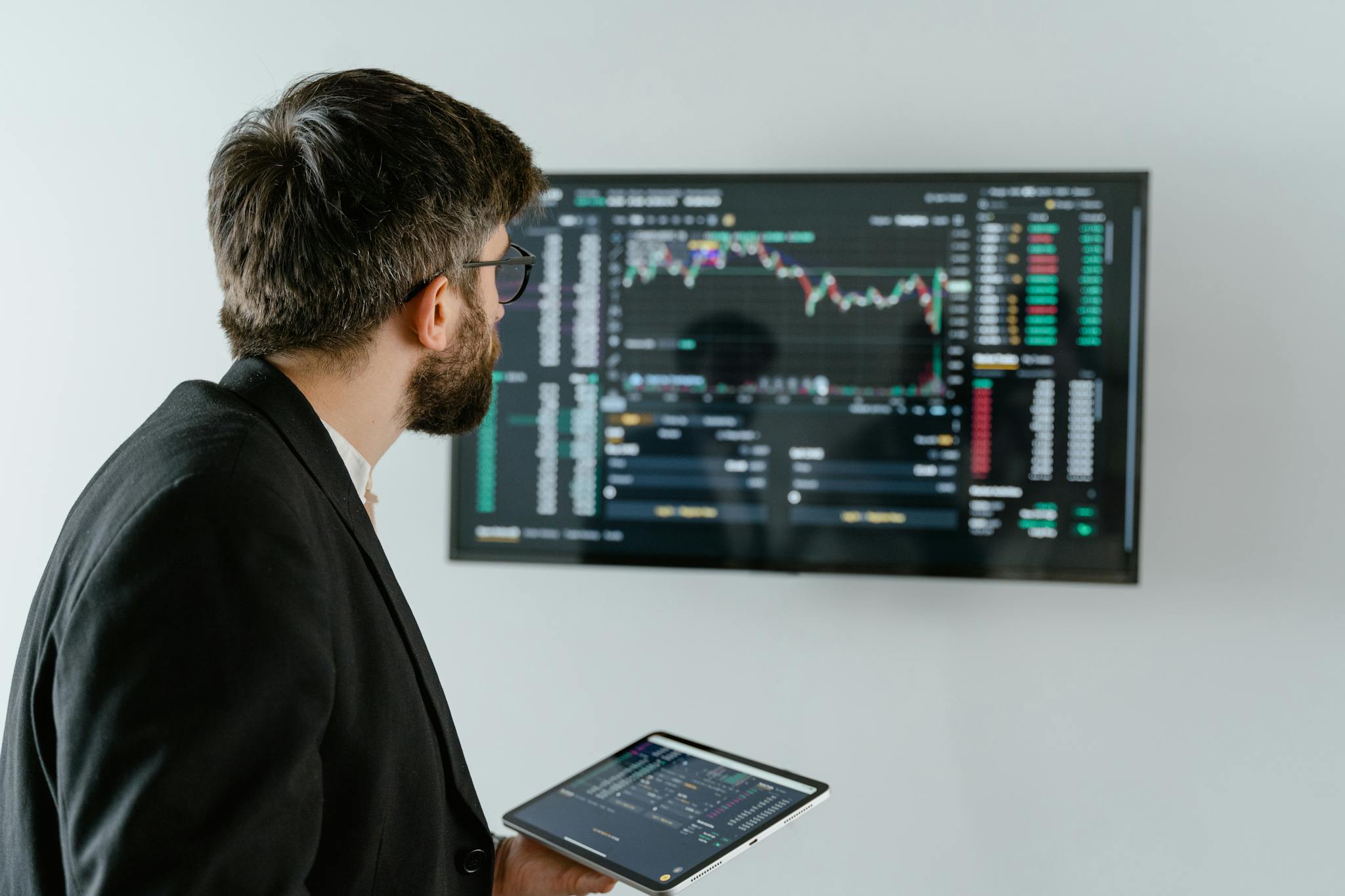 A businessman examines stock market data displayed on a monitor, holding a tablet.